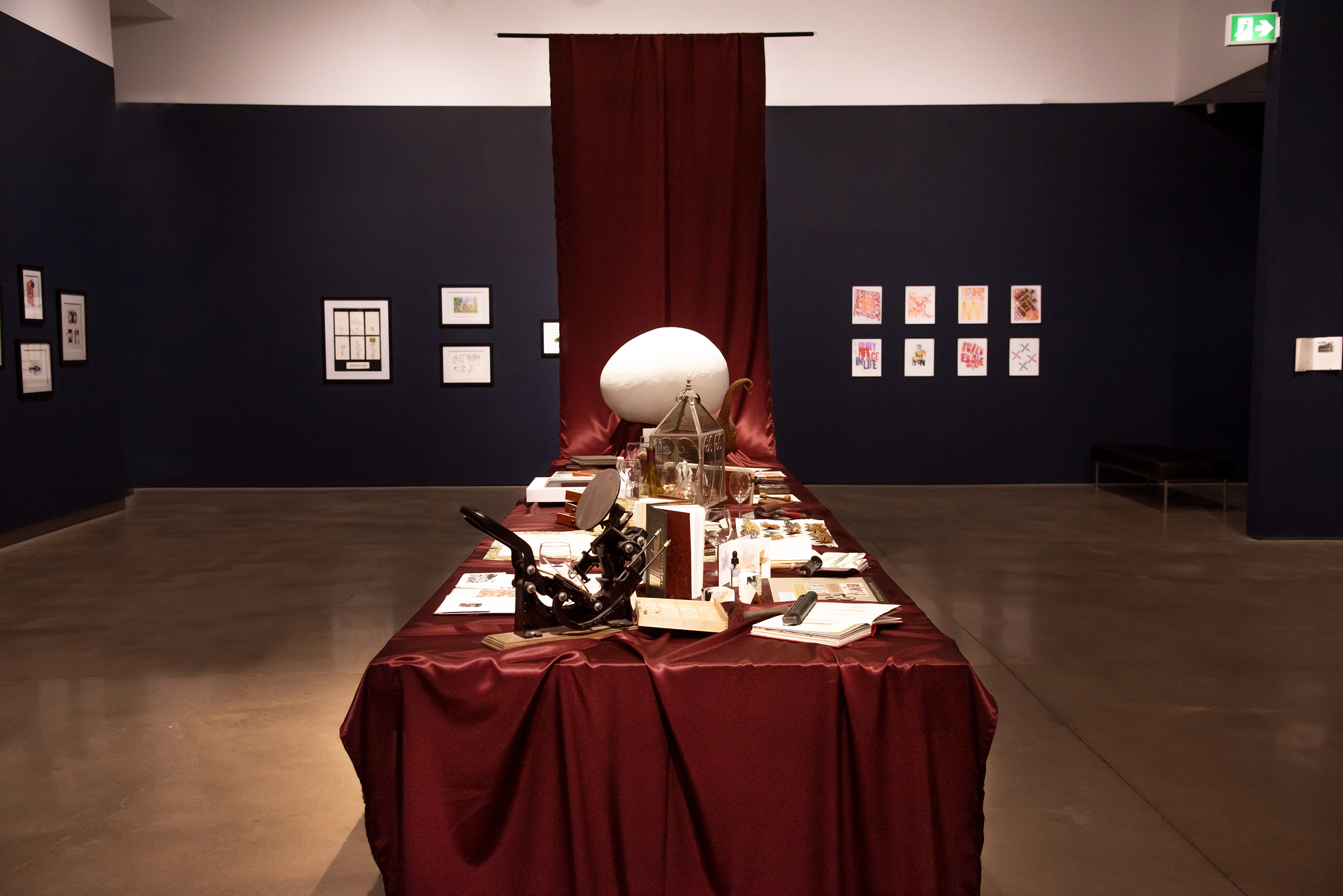 A banquet table draped in crimson satin which rises to the ceiling at one end. Atop the table are various printmaking and book art work and ephemera