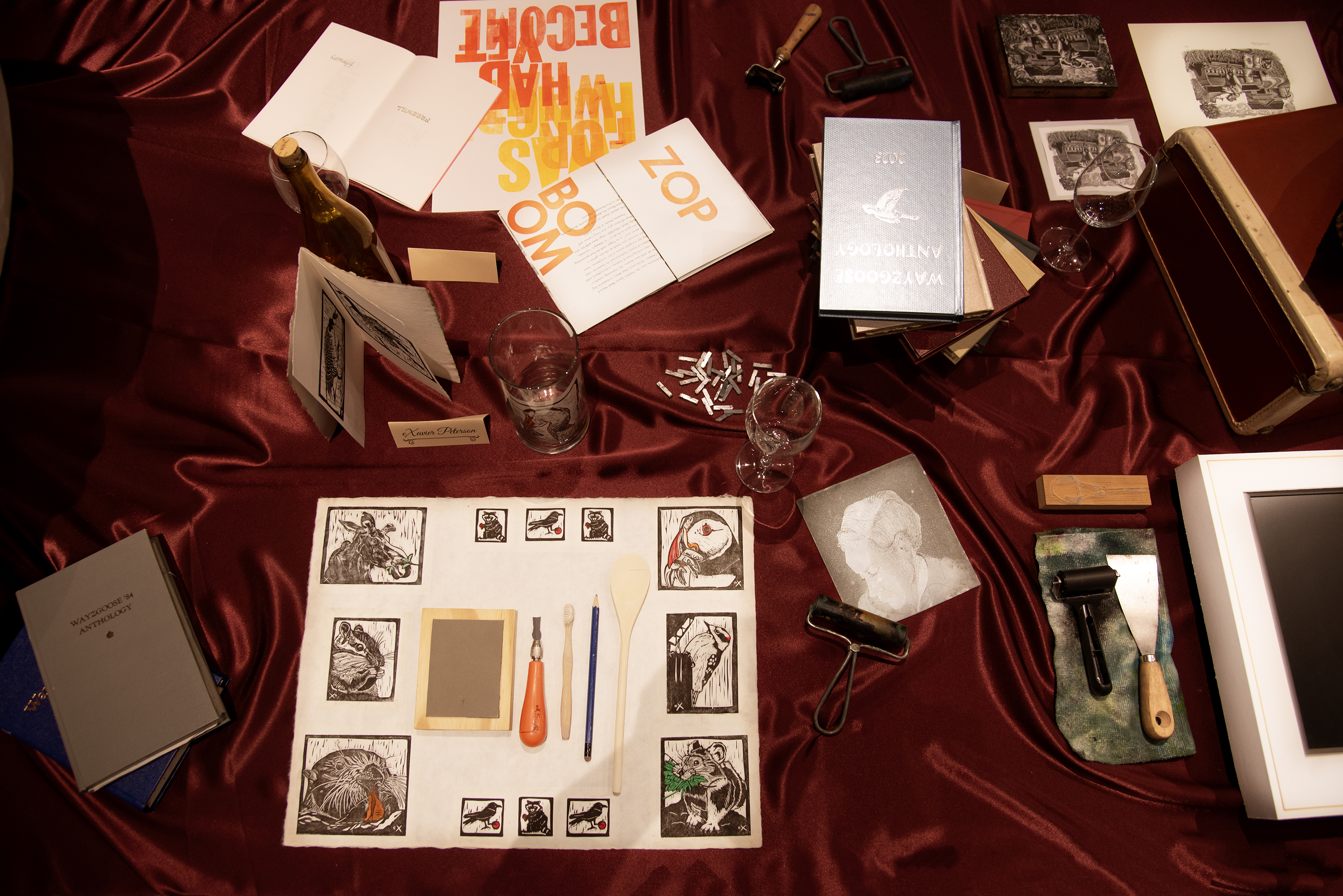 top down view of a banquet table covered in crimson satin and various printmaking and book art ephemera atop it set like table settings